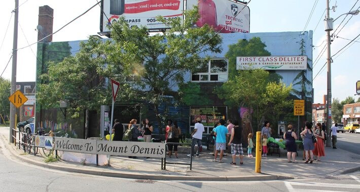 Pedestrians on a street corner with a sign reading Welcome to Mount Dennis