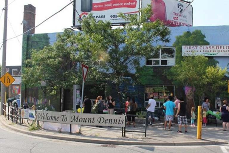 Pedestrians on a street corner with a sign reading Welcome to Mount Dennis